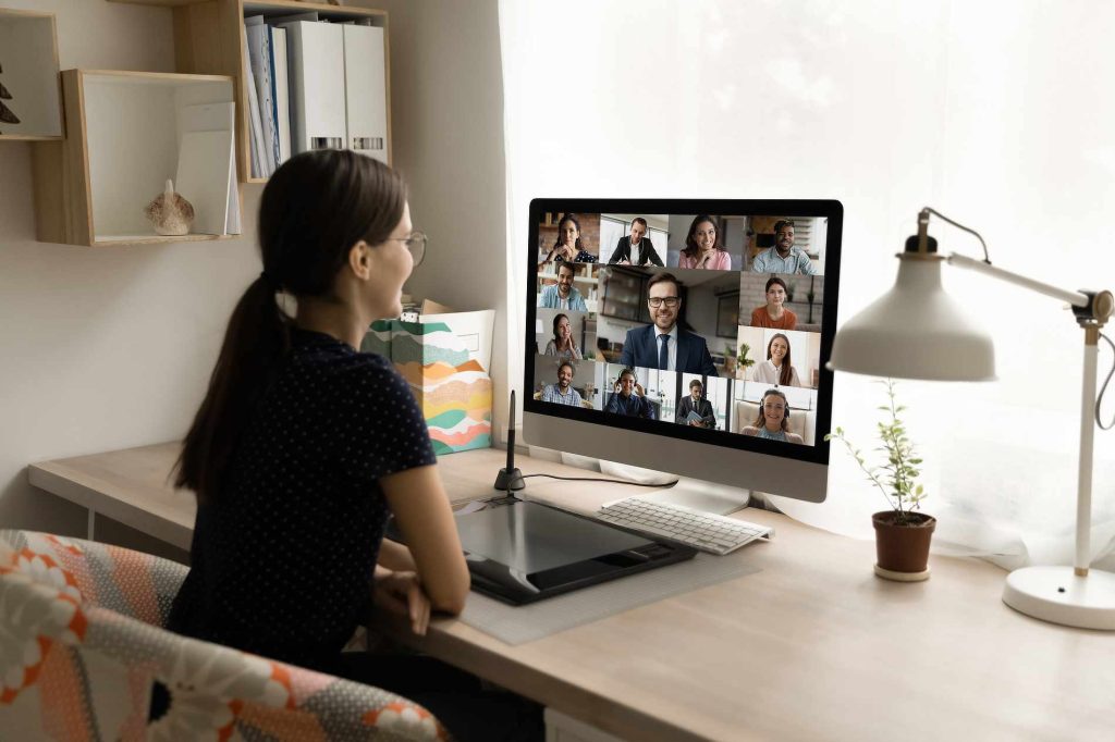 A woman with long dark hair in a ponytail sits at a desk, engaged in a video conference with twelve people on her monitor. The desk features a tablet, keyboard, and a potted plant. Sunlight streams through a nearby window, brightening the workspace.