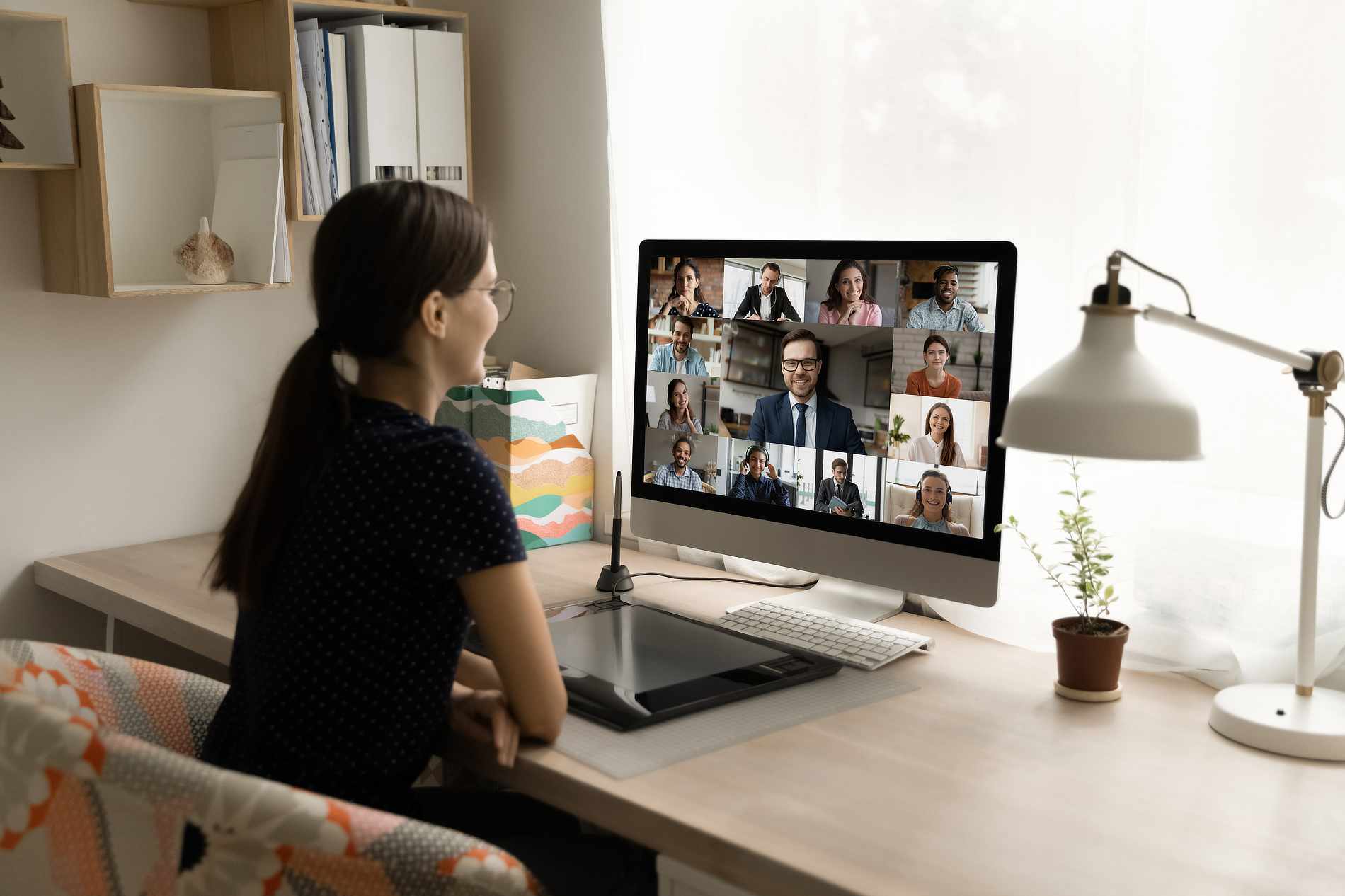A woman with long dark hair in a ponytail sits at a desk, engaged in a video conference with twelve people on her monitor. The desk features a tablet, keyboard, and a potted plant. Sunlight streams through a nearby window, brightening the workspace.