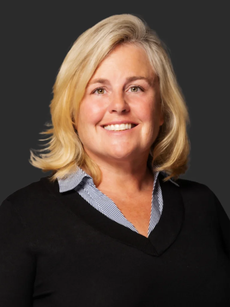 A professional headshot of Cecilia Dahl, a woman with shoulder-length blonde hair and a bright smile. She is wearing a black V-neck sweater over a collared shirt with blue and white stripes. The background is a plain dark gray, highlighting her friendly and approachable demeanor.