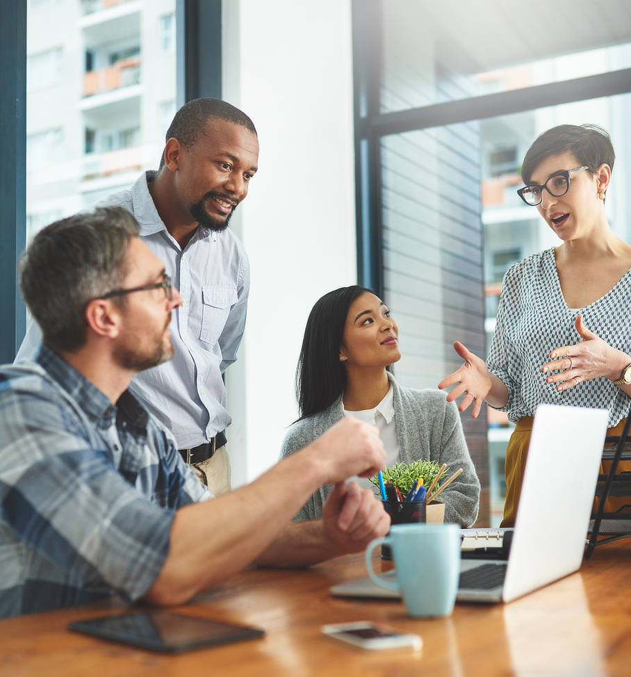 A diverse group of colleagues engaged in a discussion around a table with a laptop and tablet, symbolizing teamwork and collaborative brainstorming in a modern office environment.