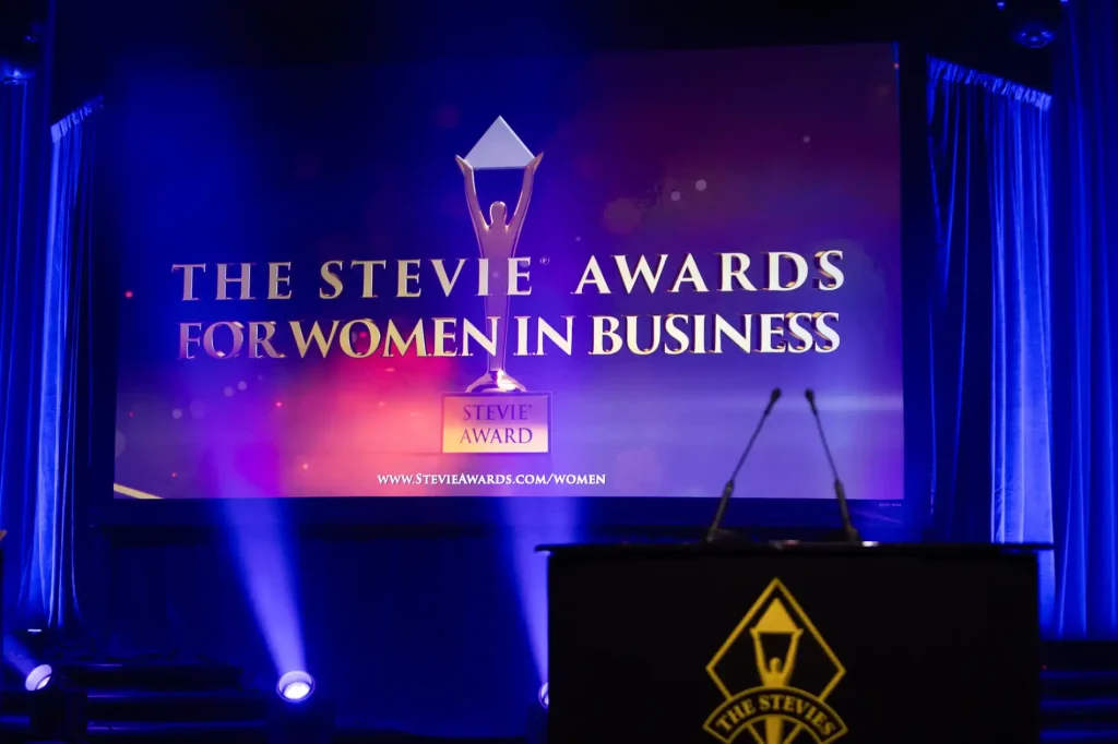 "The Stevie Awards for Women in Business" with the Stevie Awards logo and a podium in front, surrounded by blue lighting.
