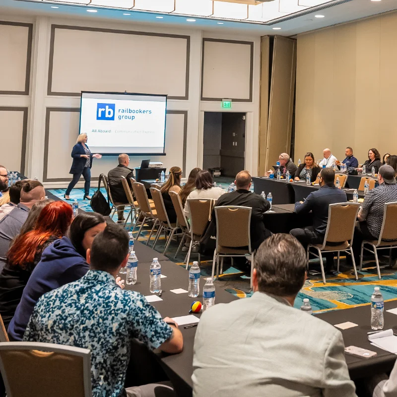 Cecilia Dahl delivers a presentation for The Remote Leadership Lab in a conference room. She walks confidently near a screen displaying the "Railbookers Group" logo, while attendees sit around tables, listening attentively.