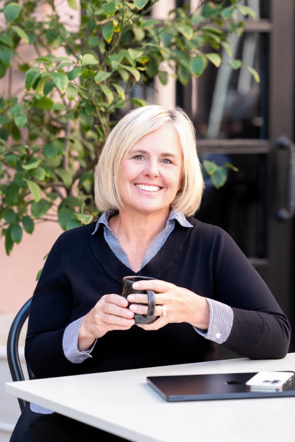 Cecilia Dahl sits at an outdoor table, smiling while holding a mug, with a laptop in front of her and greenery in the background.