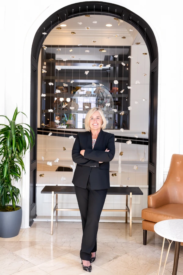Cecilia Dahl stands confidently with arms crossed in a modern office space, wearing a black suit and smiling beneath an arched doorway with hanging sculptural elements.