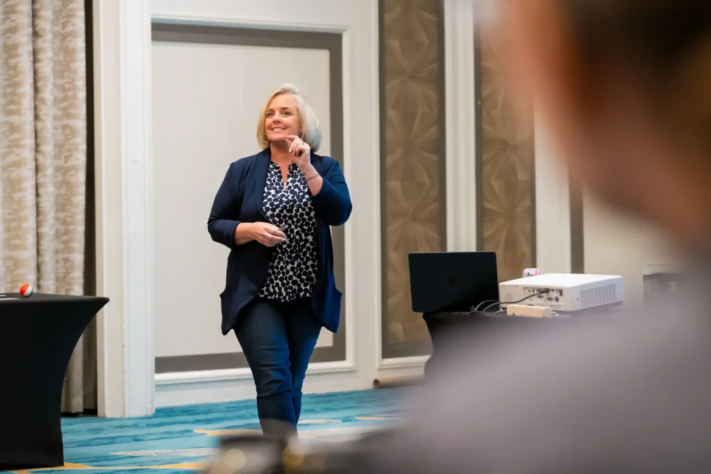 Cecilia Dahl presents to an audience in a conference room, smiling and gesturing while standing near a projector and laptop.